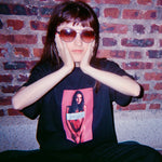 Person wearing a black t-shirt with a graphic design, sitting against a brick wall.