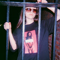 Person wearing sunglasses and a black t-shirt with a graphic design, standing behind bars.